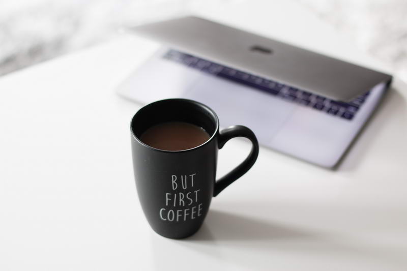 Ein Kaffee und ein Laptop im Büro. Auf der Kaffeetasse ist die Aufschrift "BUT FIRST COFFEE".